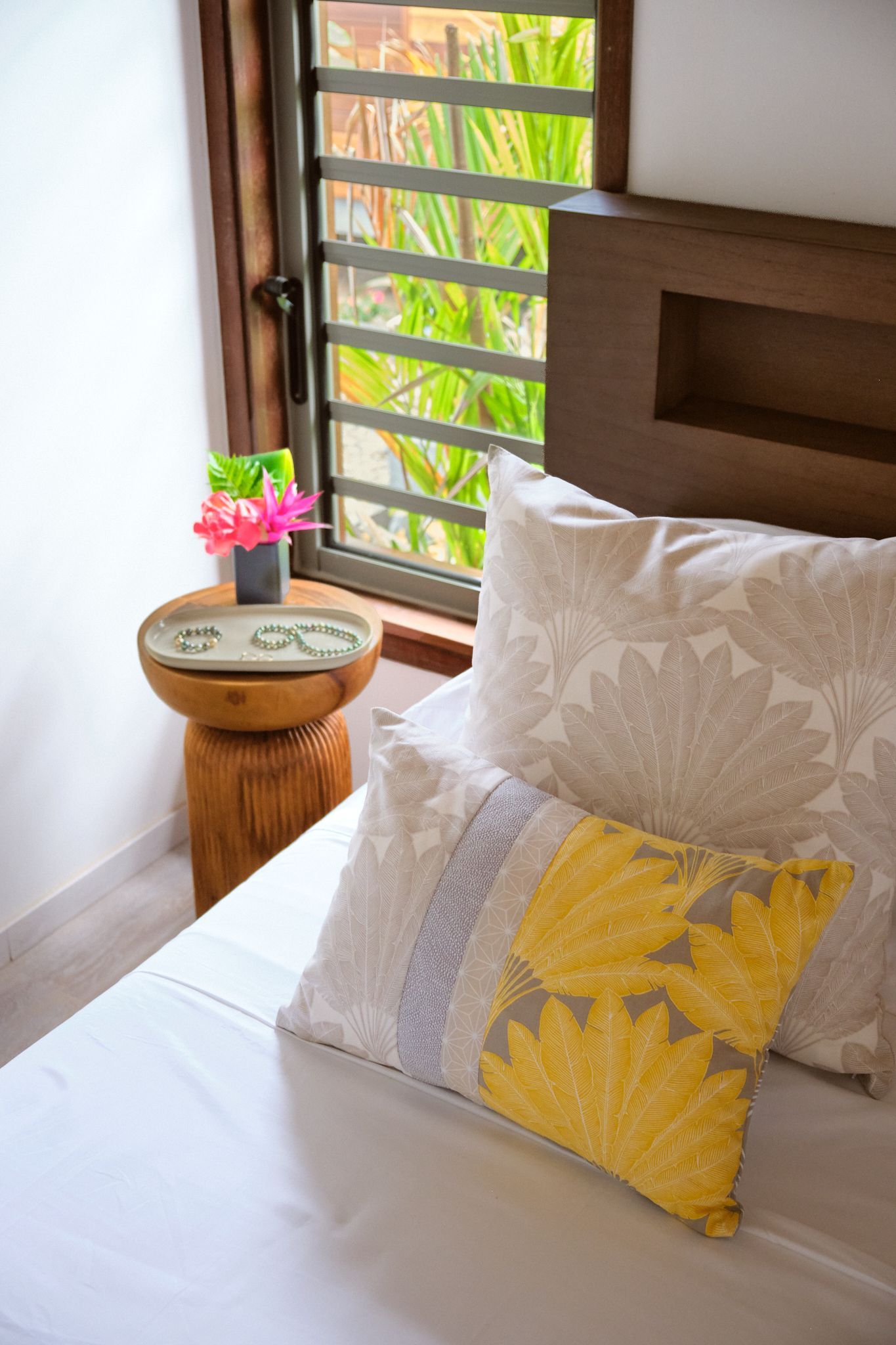 Bedroom with white sheets, patterned pillows, wooden nightstand with flowers next to a window.