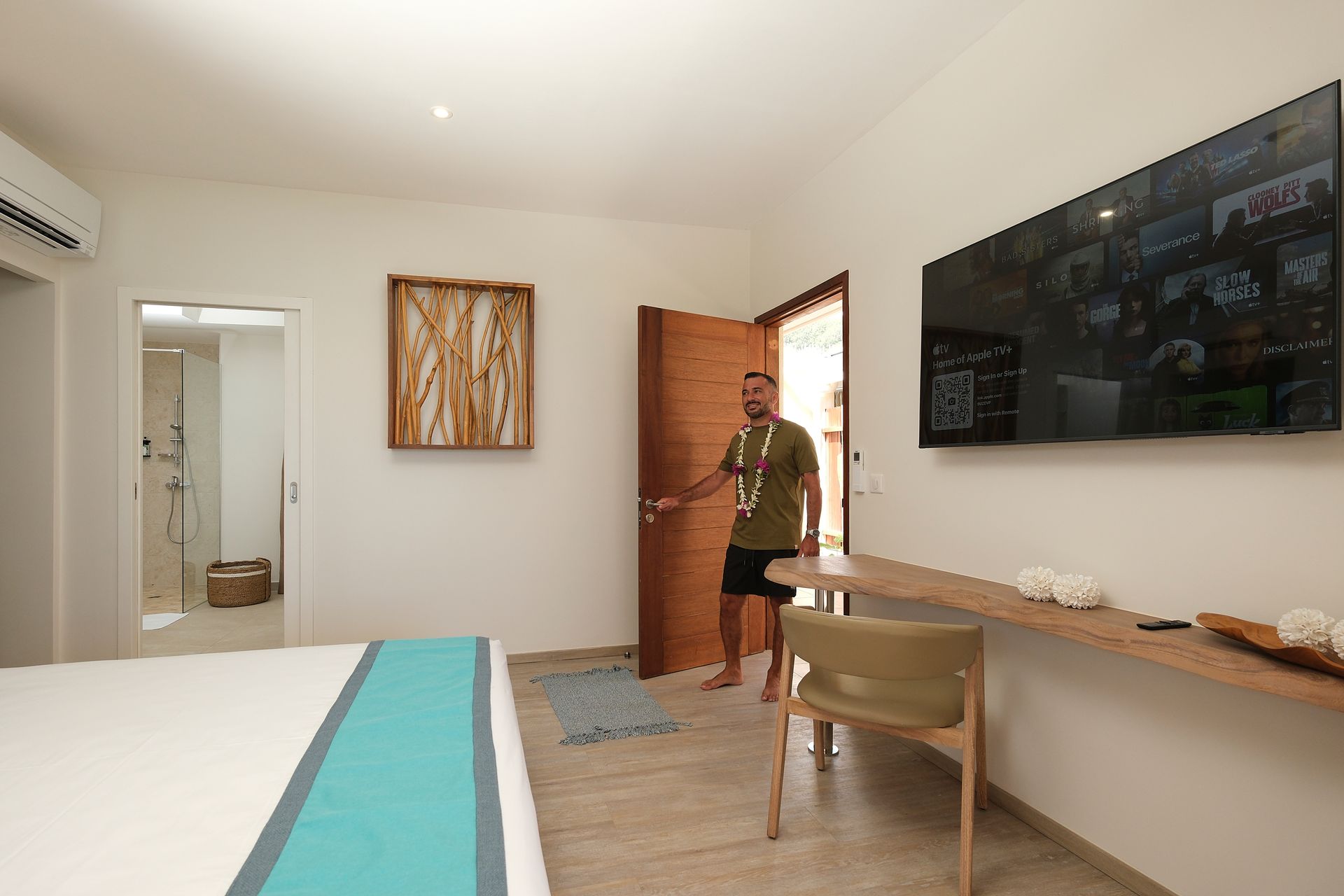Man entering a bright hotel room with a bed, TV, and open door to the outside.