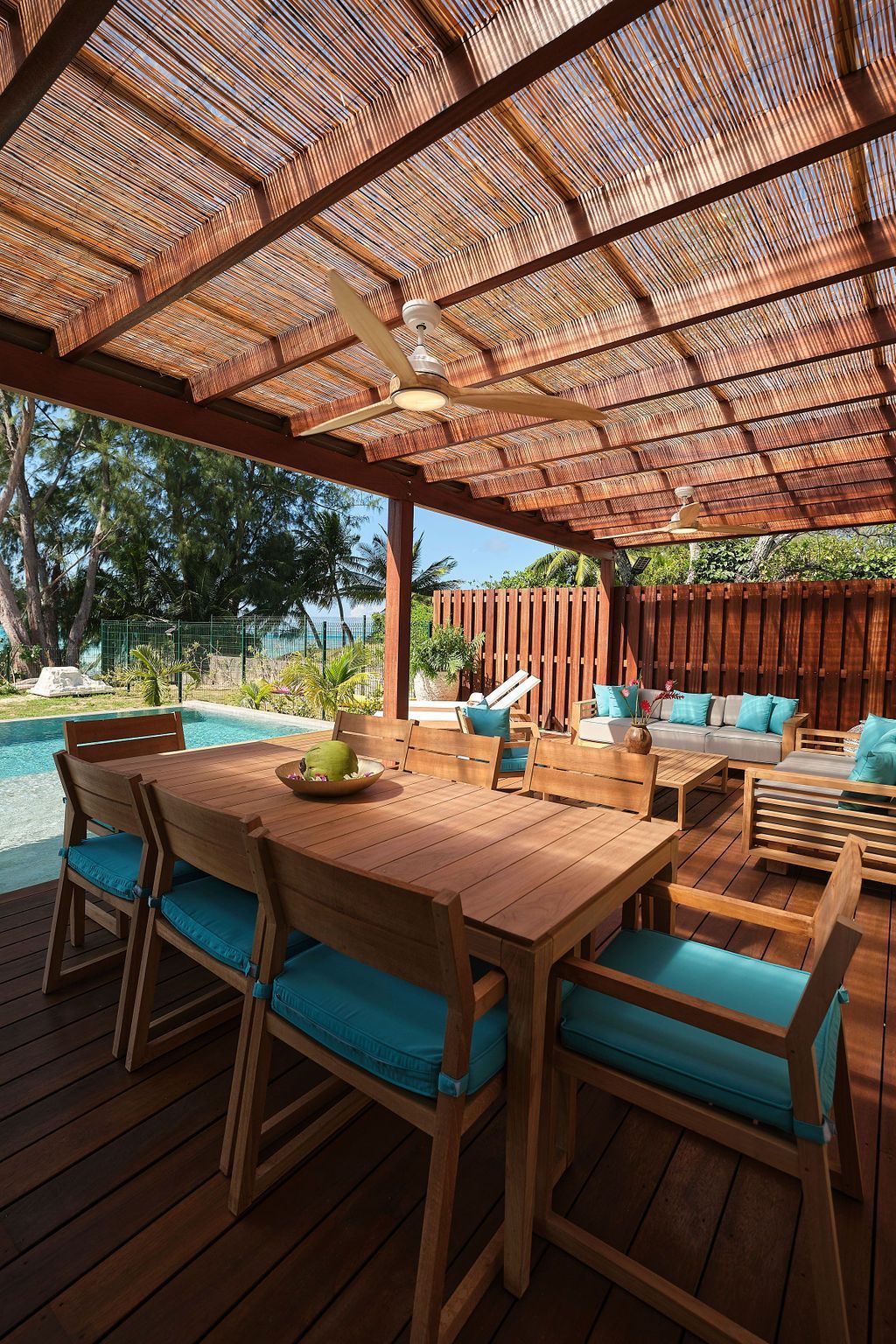 Outdoor dining area with wooden table and chairs, turquoise cushions, under a pergola roof, near a pool.