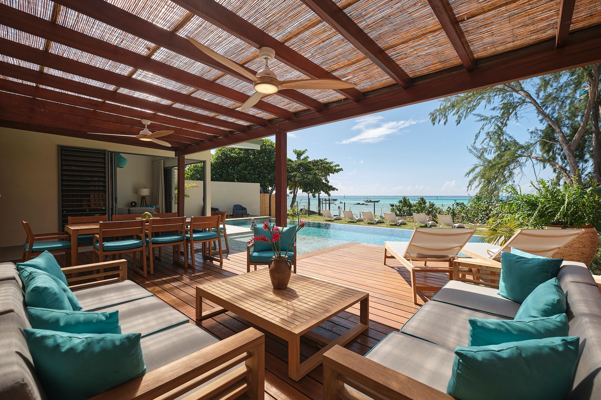 Outdoor patio with pool and ocean view; wooden furniture, blue cushions, sunny sky.