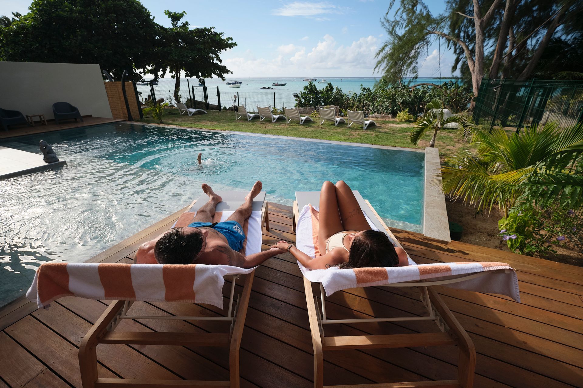 Couple relaxing on lounge chairs by a pool overlooking the ocean, holding hands.