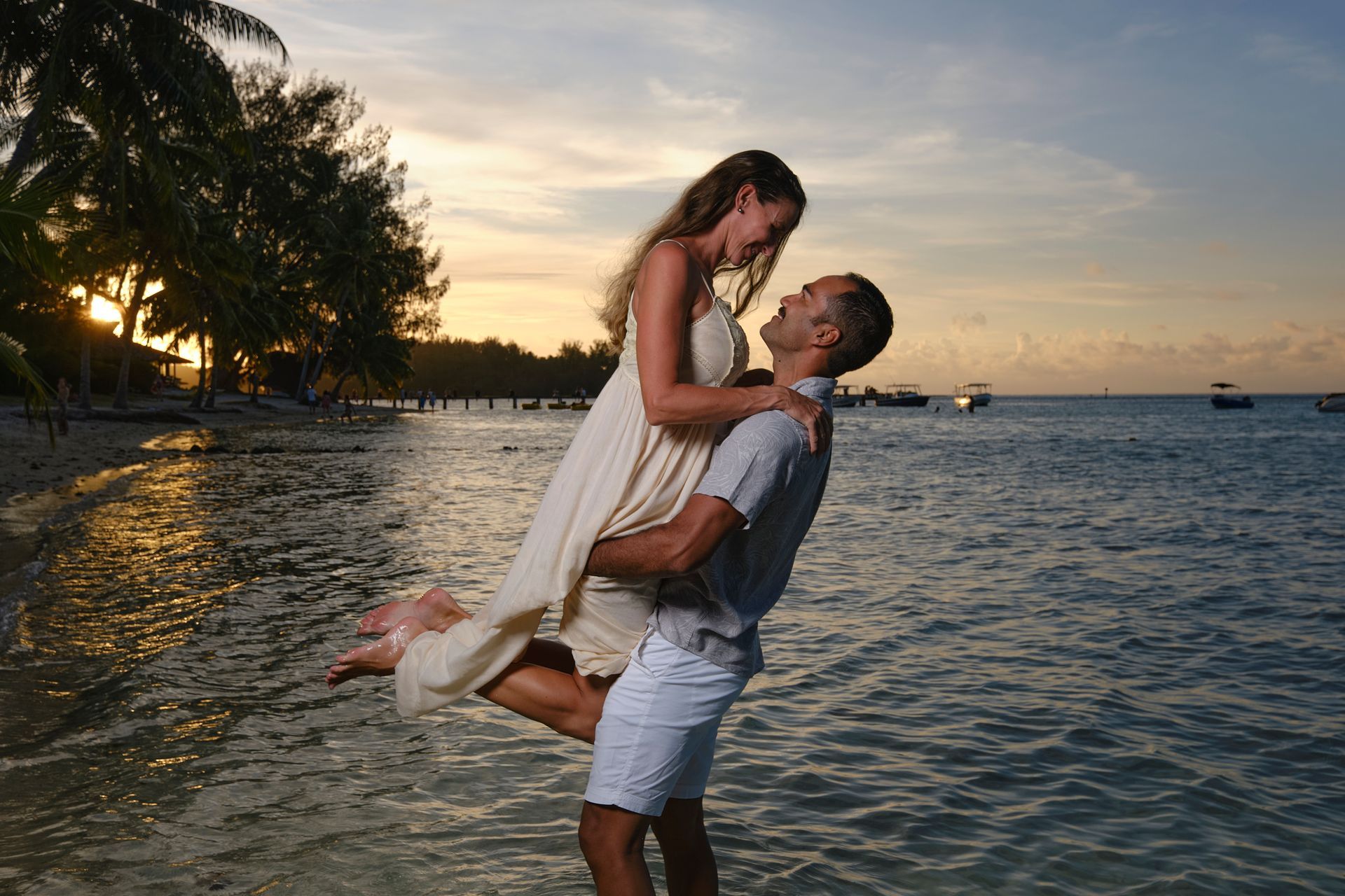Man holding woman in his arms at the beach, sunset in the background.
