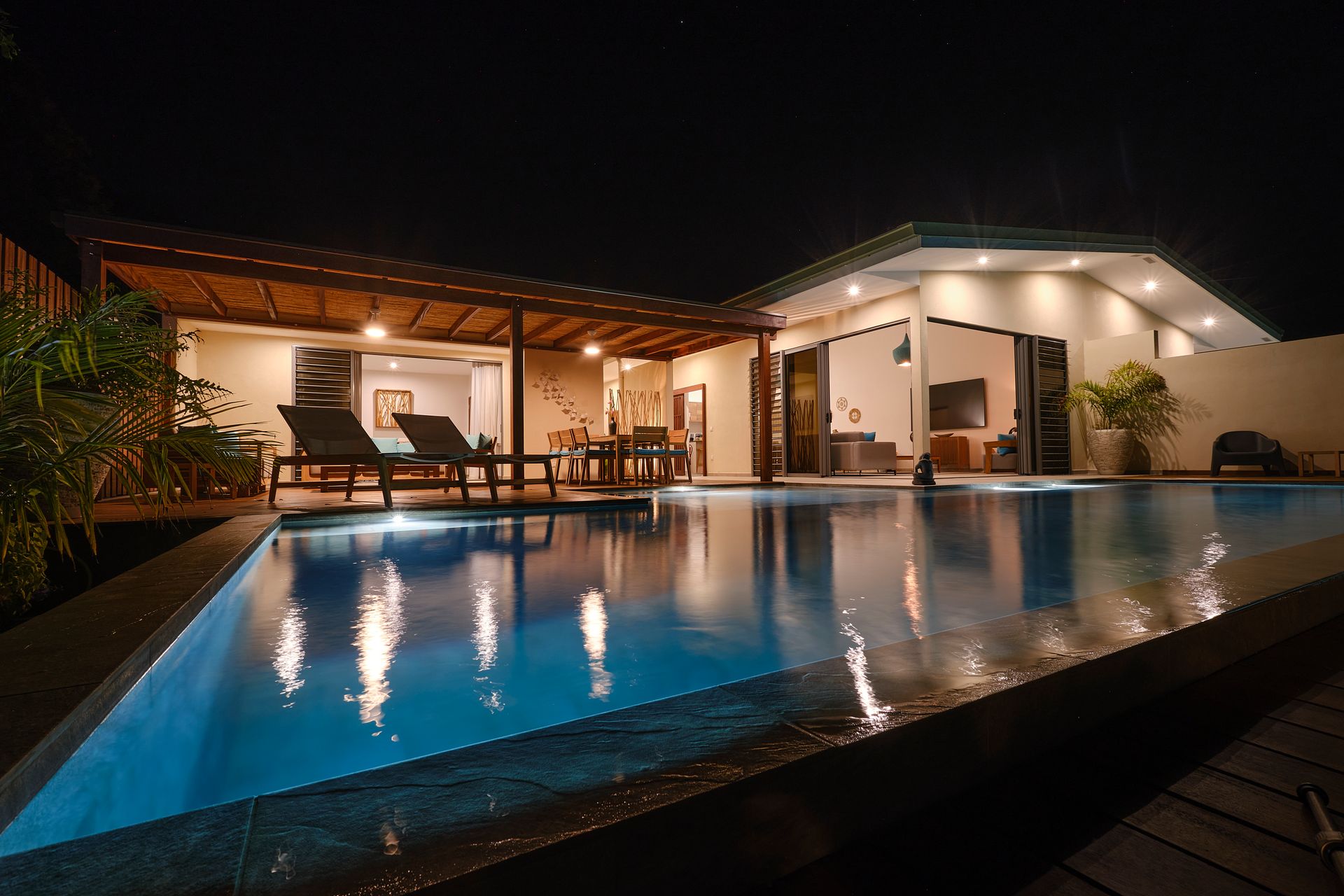 Nighttime view of a pool with reflecting lights, villa with open doors, and outdoor seating.