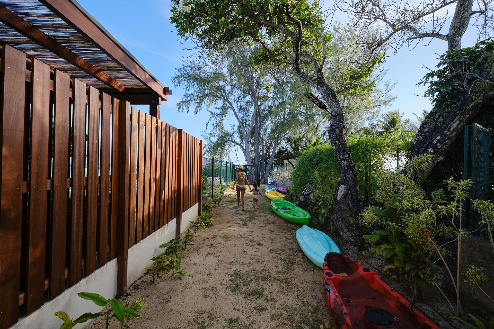 Wooden fence and path with kayaks. Two people walk towards the camera. Trees line the path.