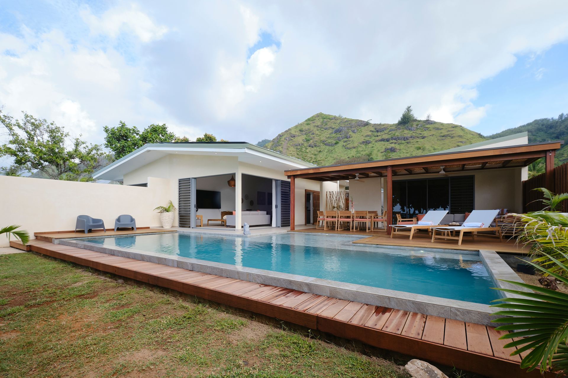 Swimming pool in front of a villa with a wooden deck, outdoor seating, and mountain in the background.