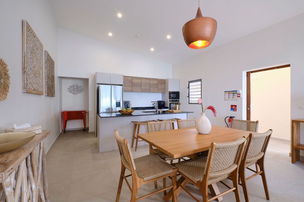 Bright dining and kitchen area with light wood table and chairs, white walls, and a copper light fixture.