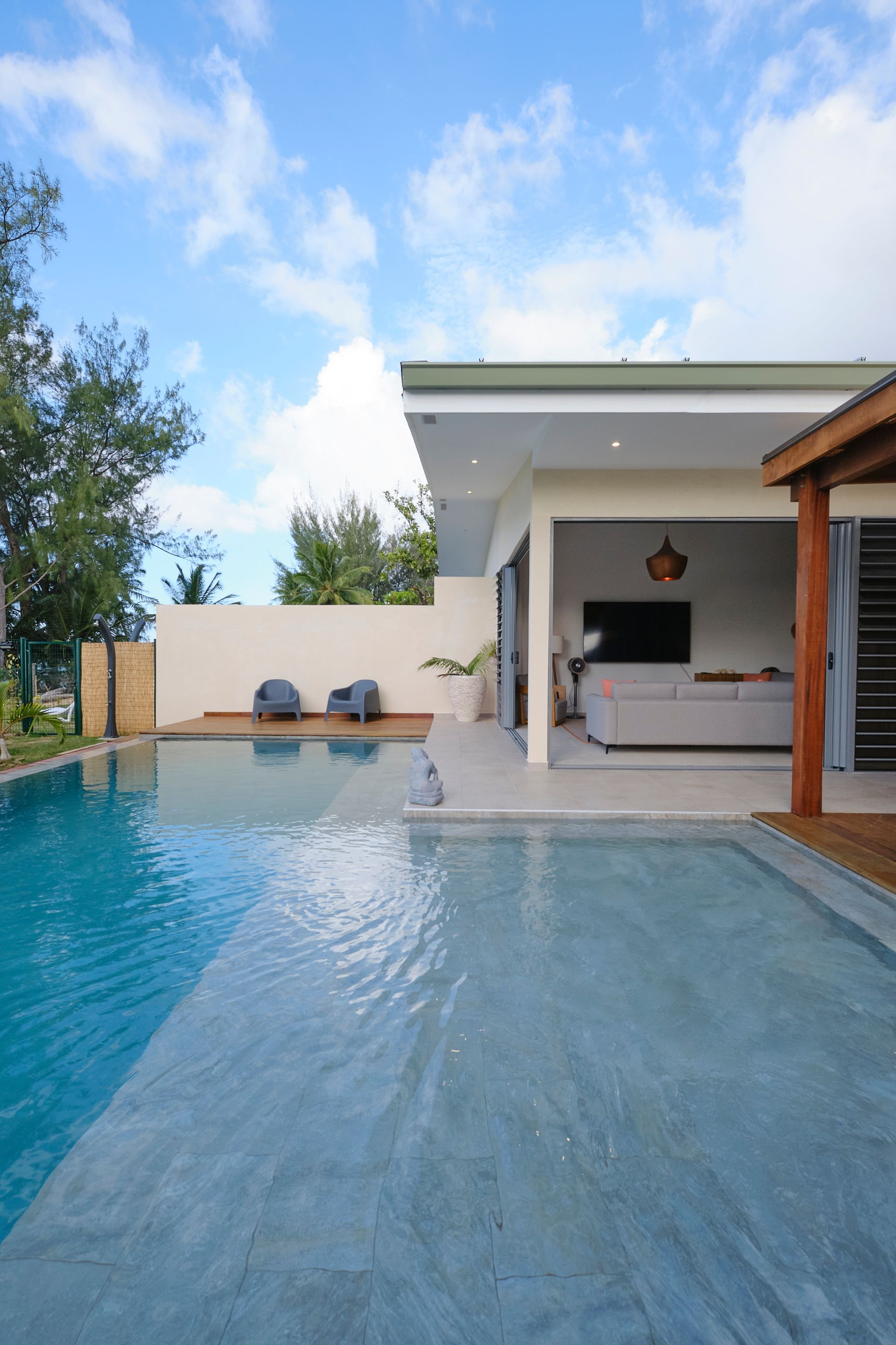Poolside view of a modern villa with a turquoise pool, patio, and partially open living area with furniture under a blue sky.