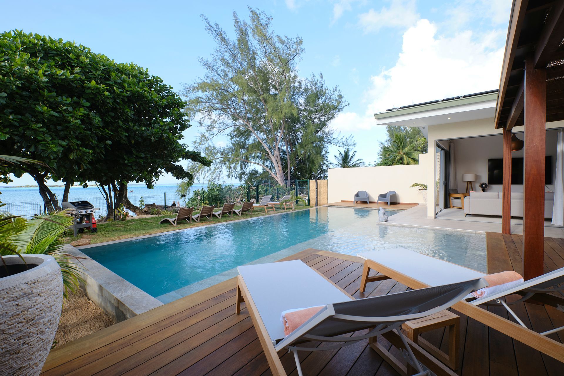 Wooden deck with pool, ocean view, and lounge chairs next to a resort building under a blue sky.