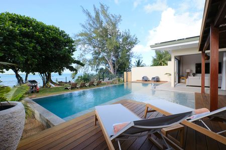 Wooden deck with pool, ocean view, and lounge chairs next to a resort building under a blue sky.