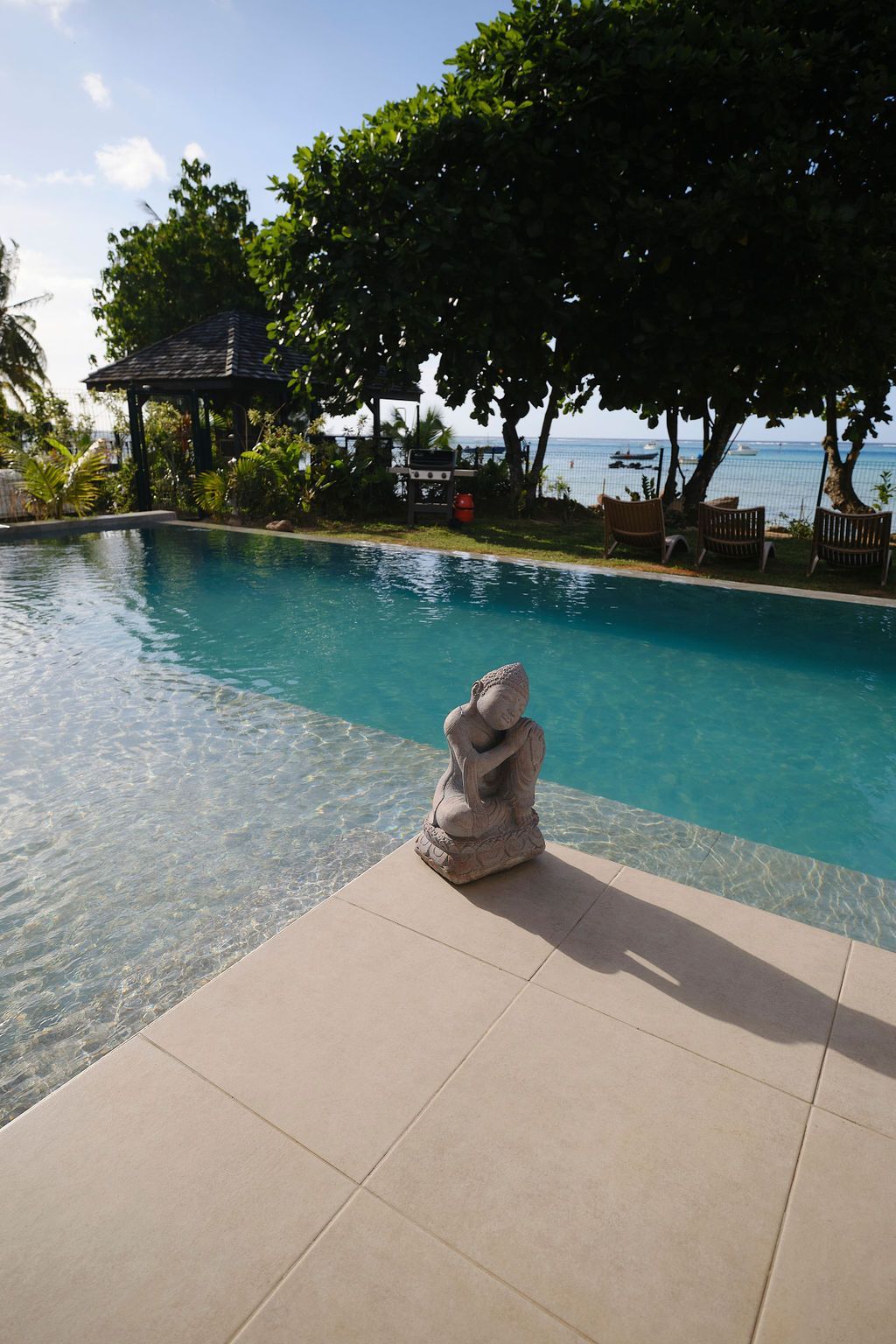 Swimming pool overlooking a beach. A stone statue sits poolside. Trees and a gazebo in the background.