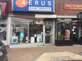 Storefronts on a sidewalk, including an AERUS Electrolux shop with blue signage.