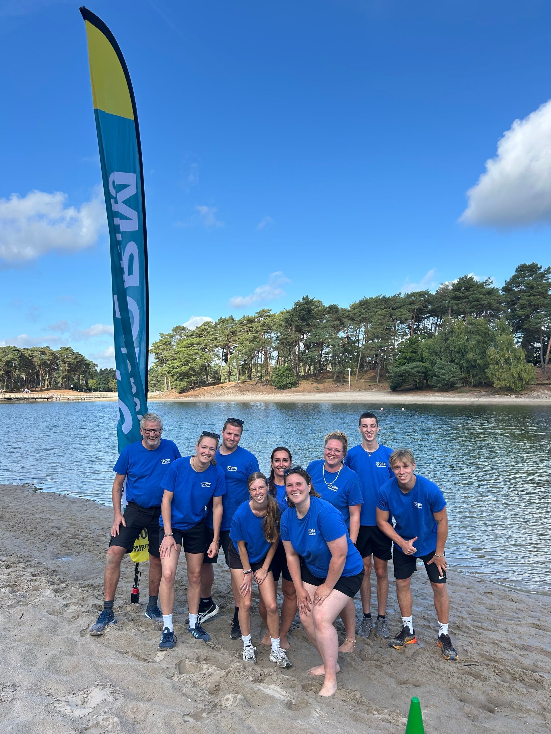 Het team poseert buiten bij een hoge blauwe evenementvlag, lachend en met hun armen omhoog op een grasveld.