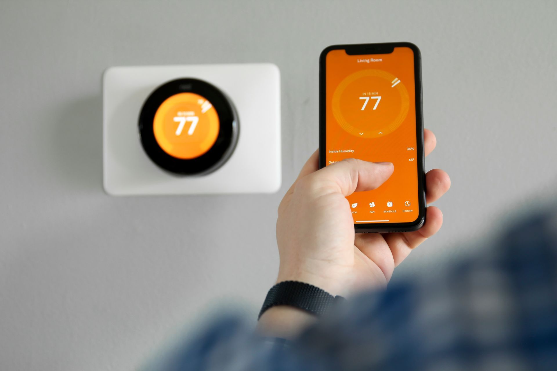 A hand holding a smartphone displays a Nest thermostat app, matching the 77-degree temperature on a wall-mounted unit.
