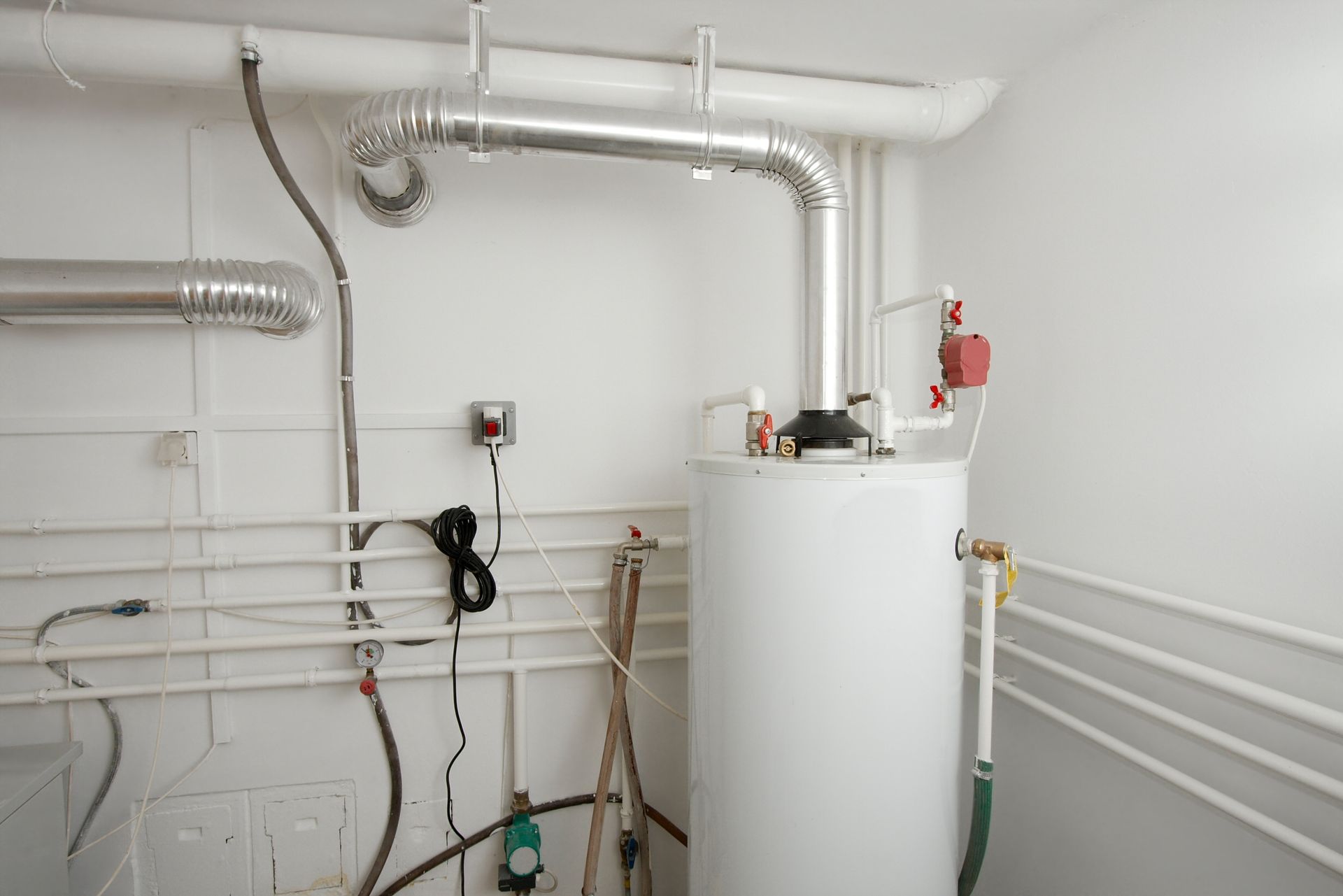 A white water heater stands in a basement room with various metal pipes and ventilation ducts along the wall.