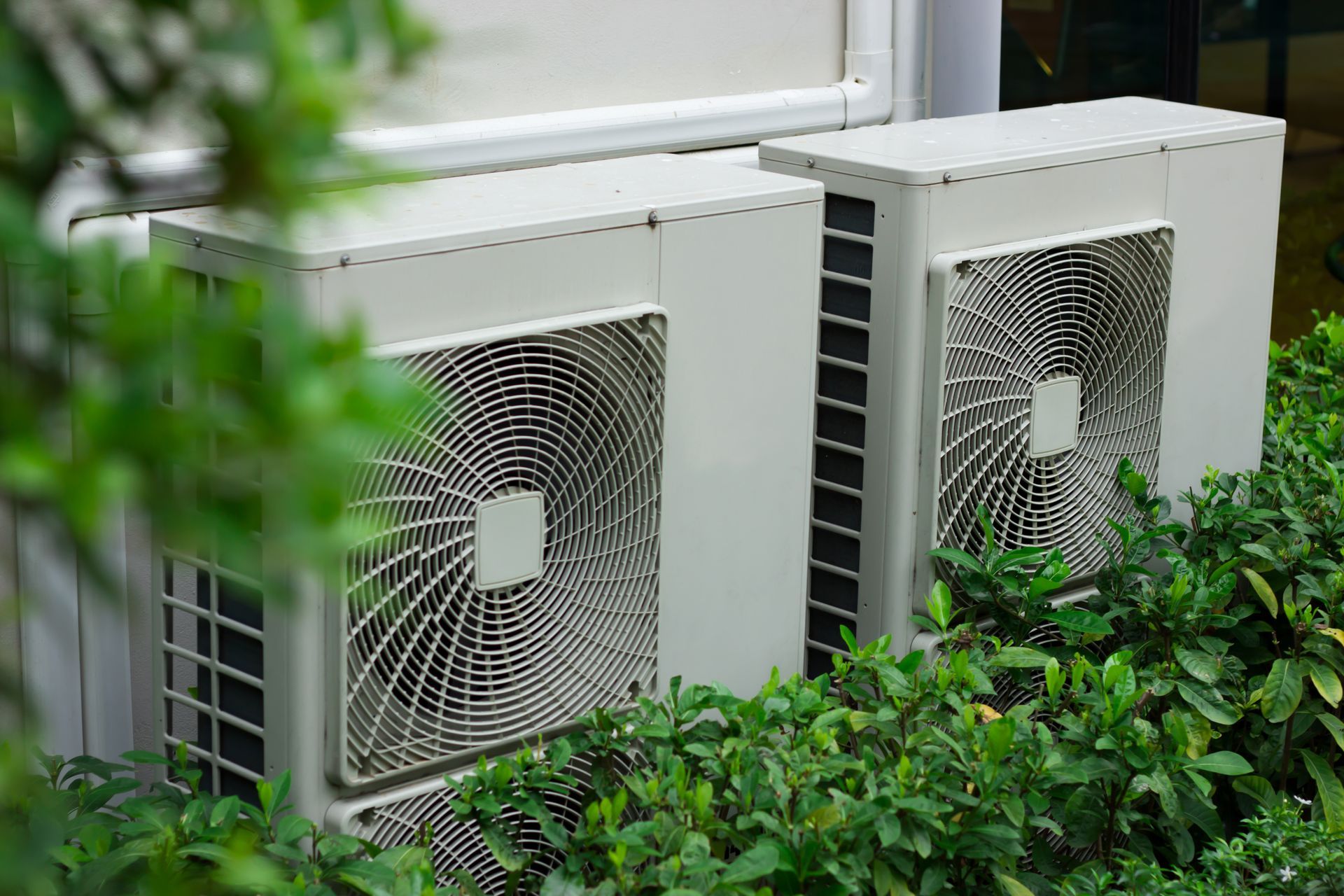Two white outdoor air conditioning units partially obscured by green leafy bushes.