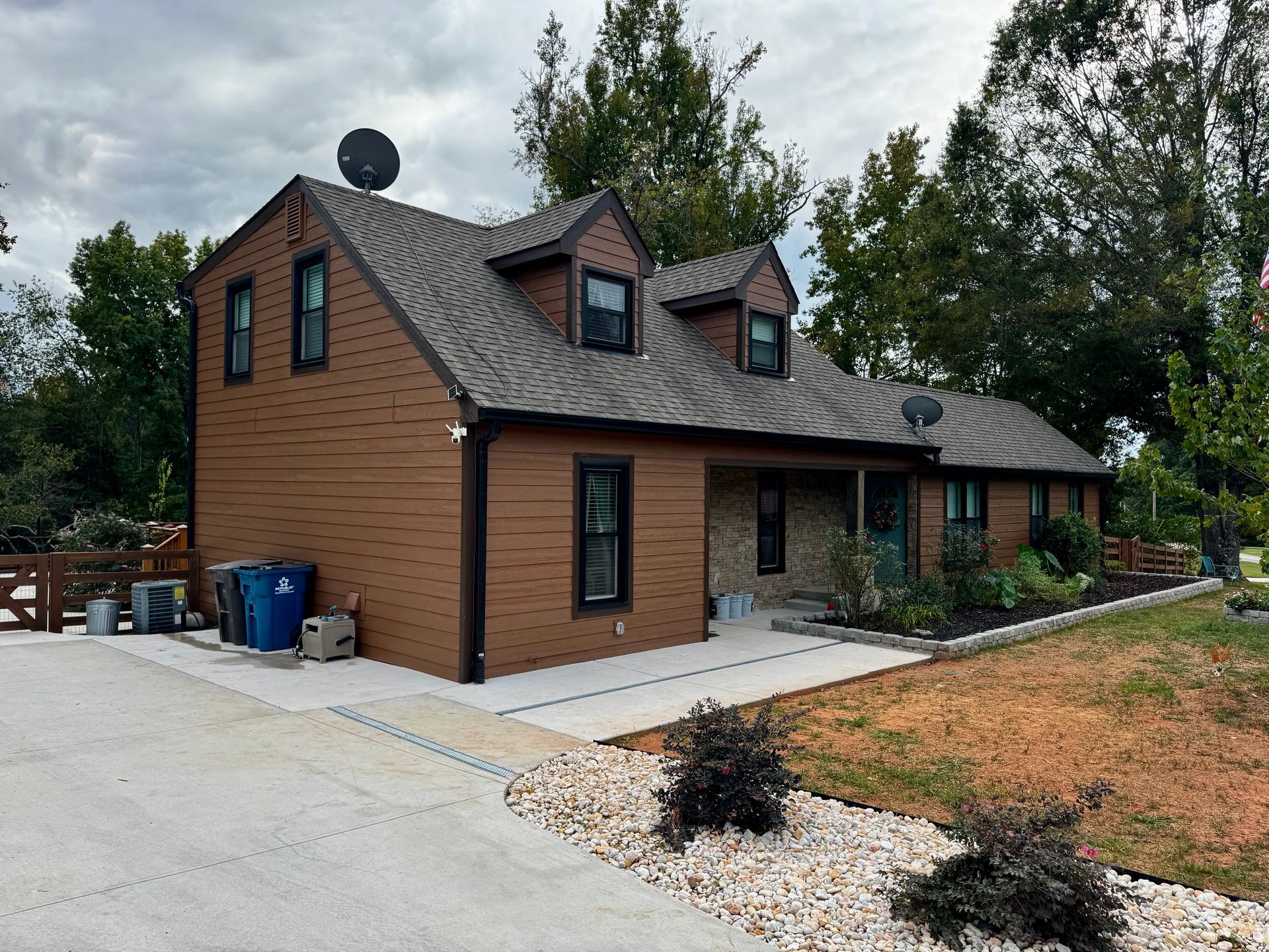 Brown house with a steep roof, stone front porch, and driveway beside a landscaped yard