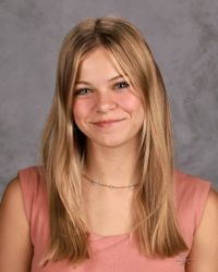 Portrait of a smiling person with straight blonde hair, wearing a pink top against a gray backdrop