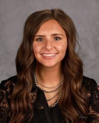 Smiling woman with long brown hair wearing a black lace top and layered necklaces against a gray backdrop