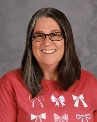 Smiling person with glasses and long dark hair wearing a red shirt with white bows against a gray background