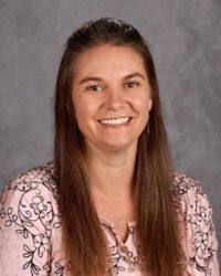 Smiling woman with long brown hair in a pink patterned blouse against a gray studio backdrop