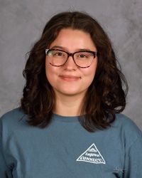 Portrait of a person with dark wavy hair and glasses, wearing a blue shirt, against a gray background