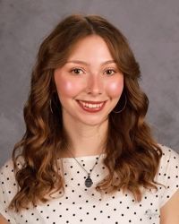 Smiling woman with wavy brown hair, white polka-dot top, and necklace against a gray background