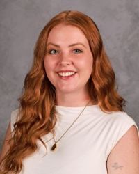 Smiling woman with long red hair wearing a white top against a gray backdrop