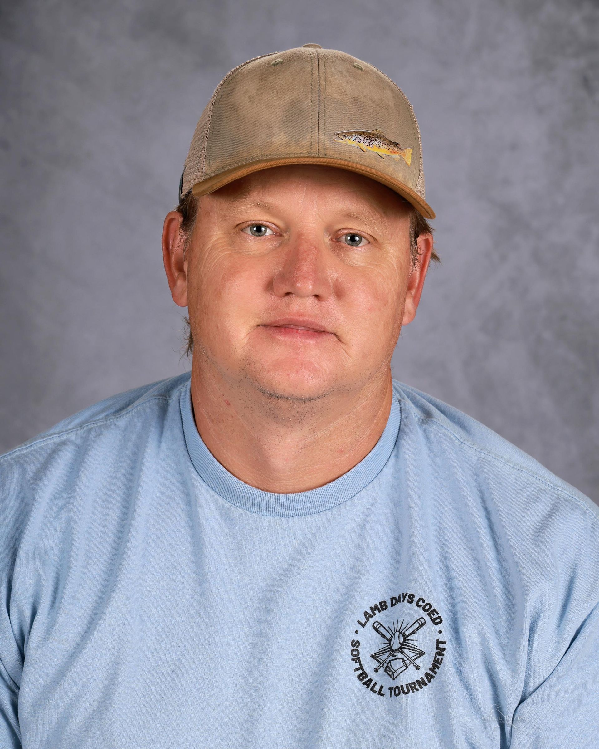 A person wearing a light blue long-sleeved shirt and a tan baseball cap, positioned against a gray studio background.