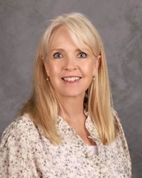 Portrait of a smiling woman with blonde hair in a floral blouse against a gray background
