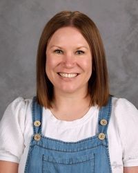 Smiling woman with shoulder-length brown hair wearing a white shirt and denim overalls against a gray backdrop