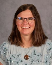 Portrait of a smiling woman with glasses, brown hair, and a patterned top against a gray background