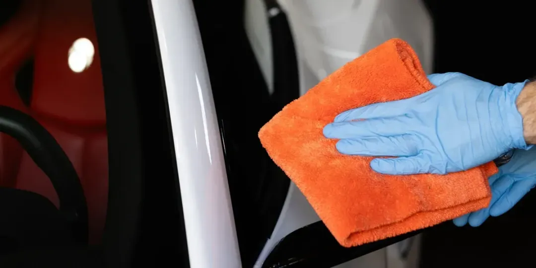 A hand in a blue glove using an orange microfiber cloth to wipe a white car surface.