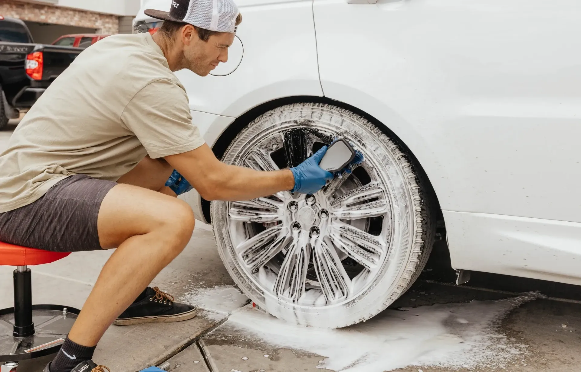 Man cleaning a car wheel with soapy water. He wears a hat, gloves, and shorts while sitting on a stool.