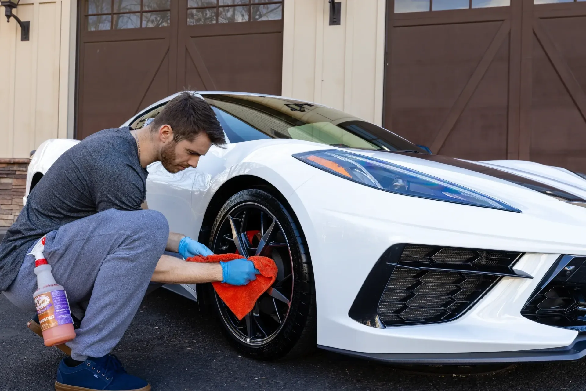 Man cleans the wheel of a white sports car with a red cloth. Spray bottle nearby. Brown garage doors in the background.
