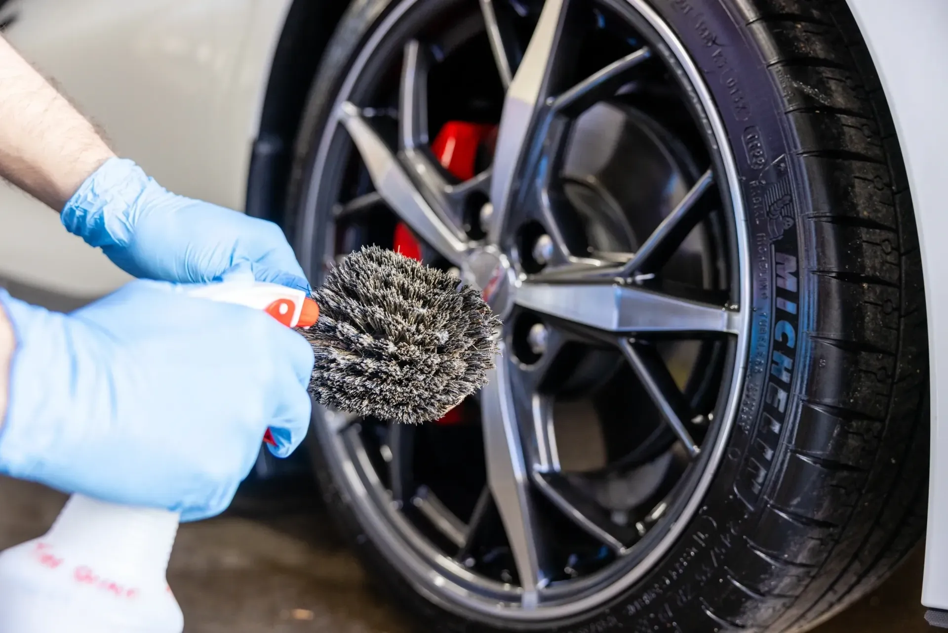 Person in blue gloves cleaning a car wheel with a brush and spray bottle.