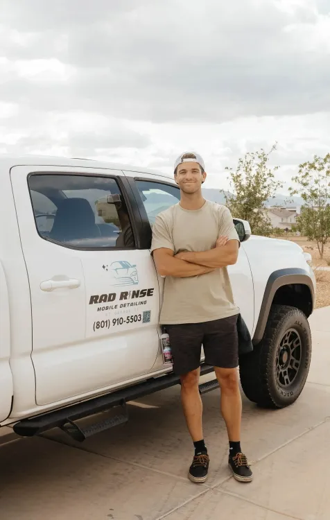 Man standing with arms crossed next to a white truck with business logo; outdoors.