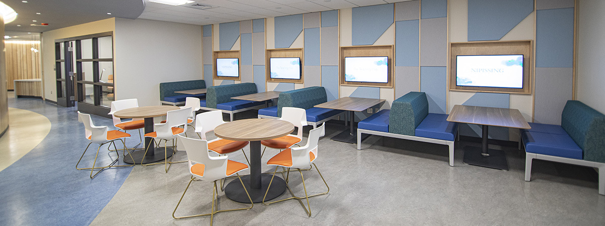 A modern waiting area with seating, tables, and screens on the walls. Light blue and orange color scheme.