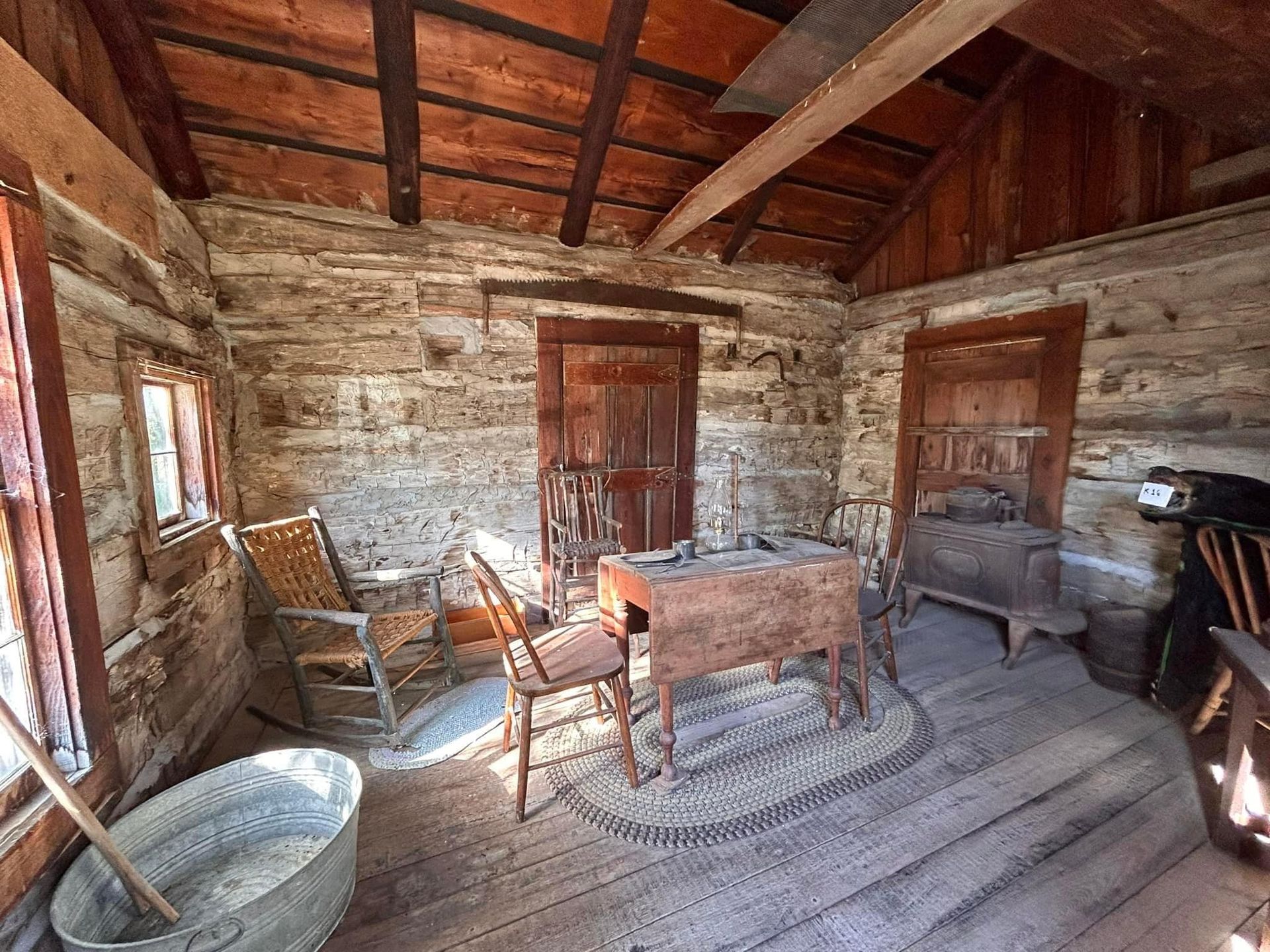 Interior of an old wooden cabin with rustic furniture, including chairs, a table, and a wood-burning stove.