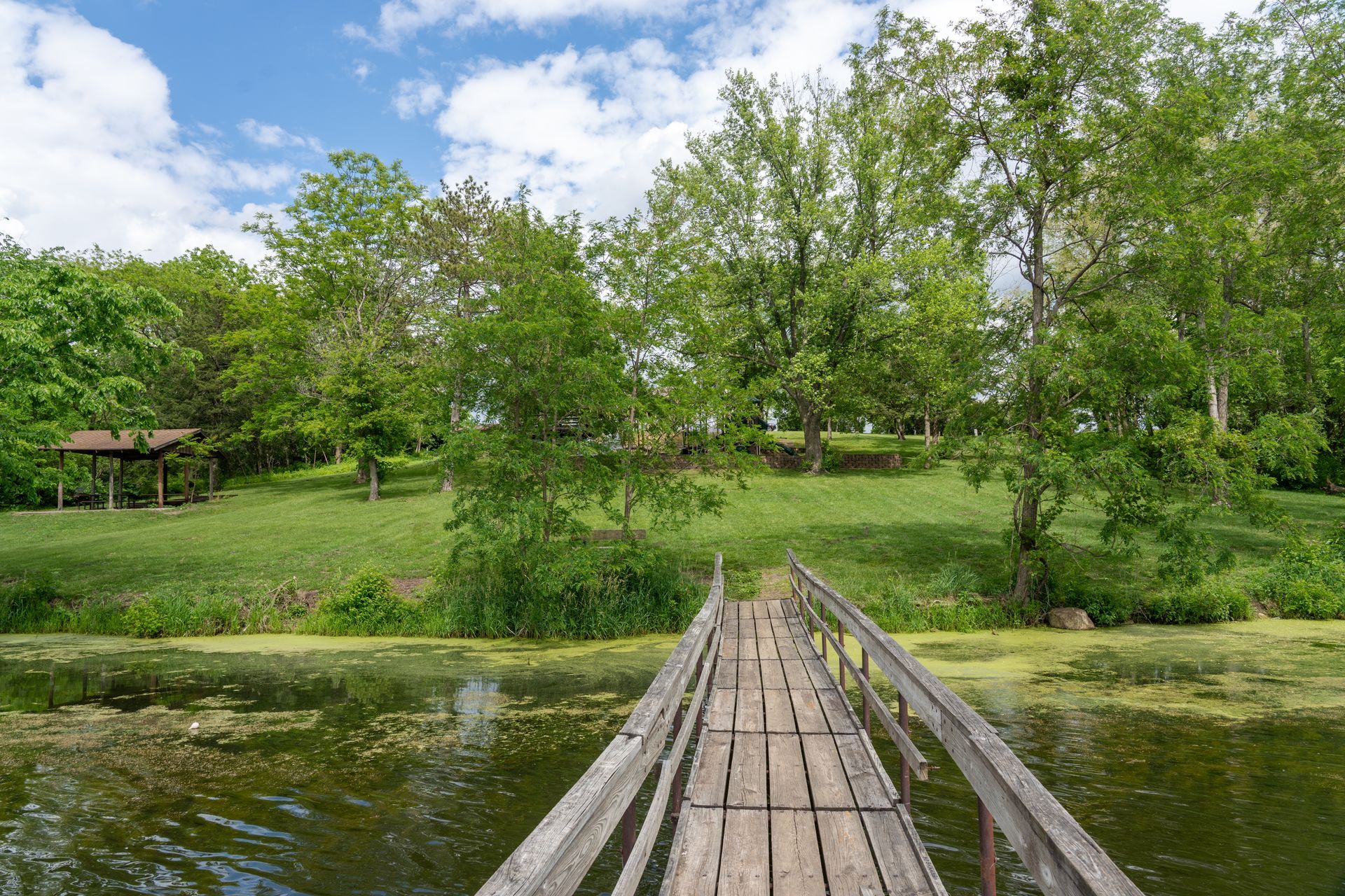 Wooden bridge over water at East Lake Park in Osceola, IA that leads to a grassy hill with trees under a blue sky.