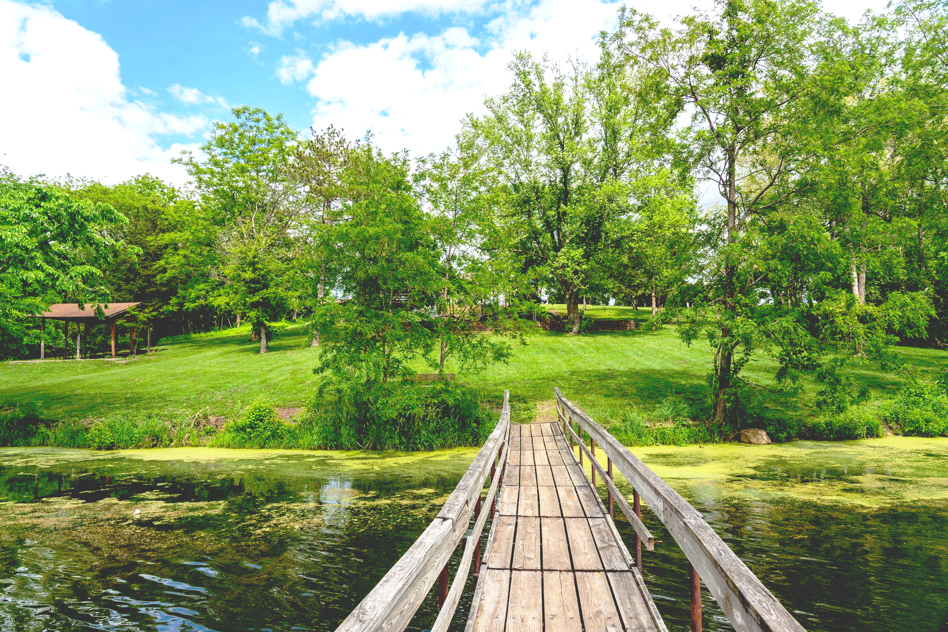Wooden bridge over East Lake in Osceola, Iowa leads to a grassy hill with trees and a gazebo under a cloudy sky.