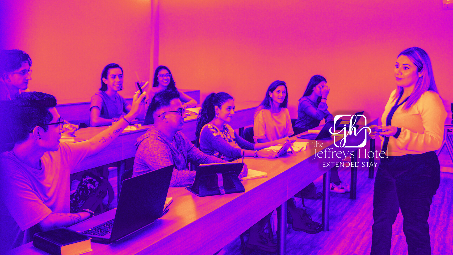 A teacher lectures students in a classroom. Students sit at desks, some using laptops. Fluorescent lighting.