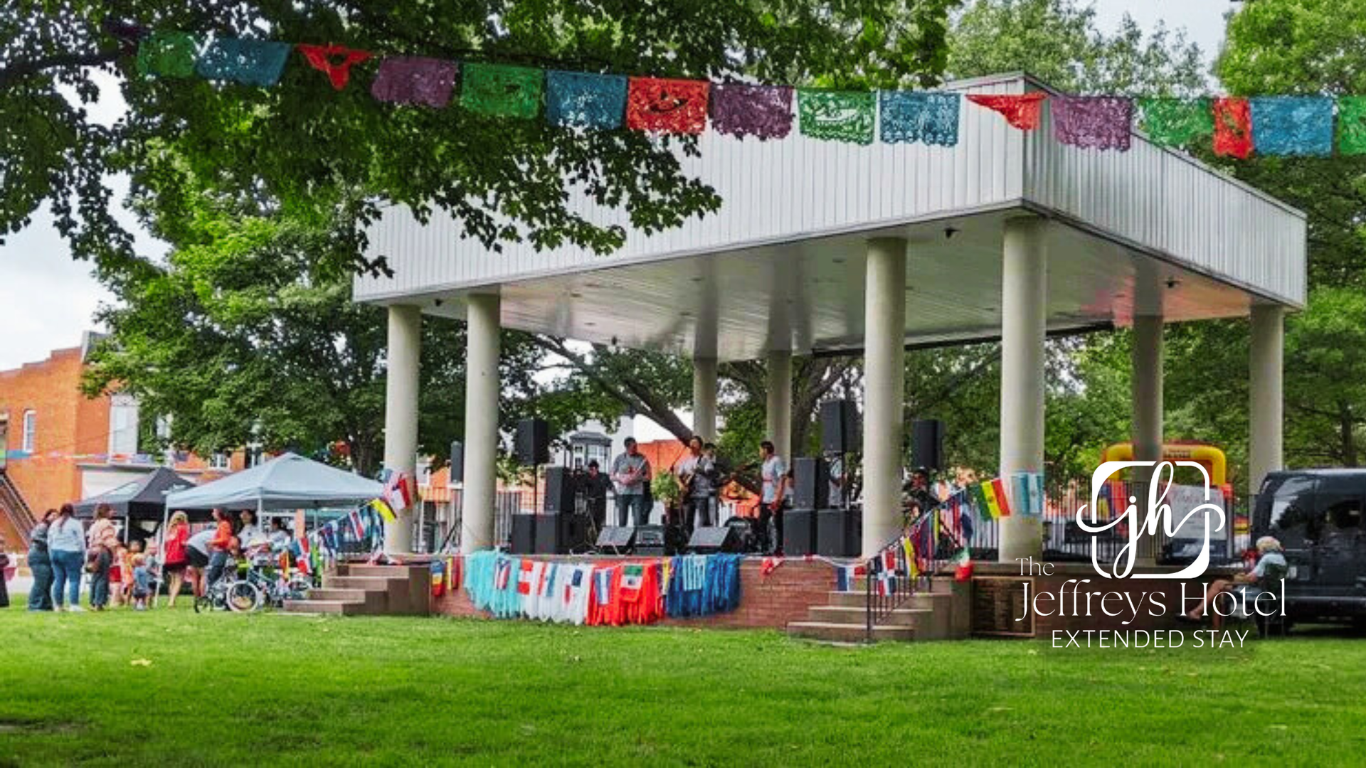 Outdoor concert at a park stage decorated with colorful flags; people gather on the grass to watch.