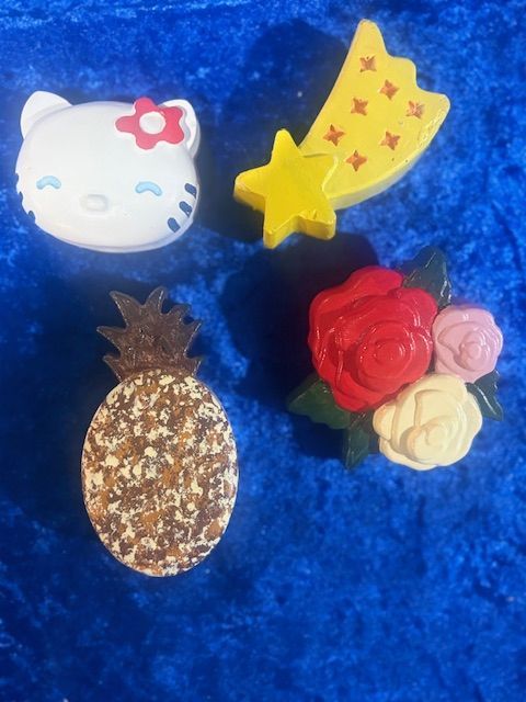 Four small decorative clips on blue fabric: Hello Kitty, yellow crescent moon, pineapple, and roses