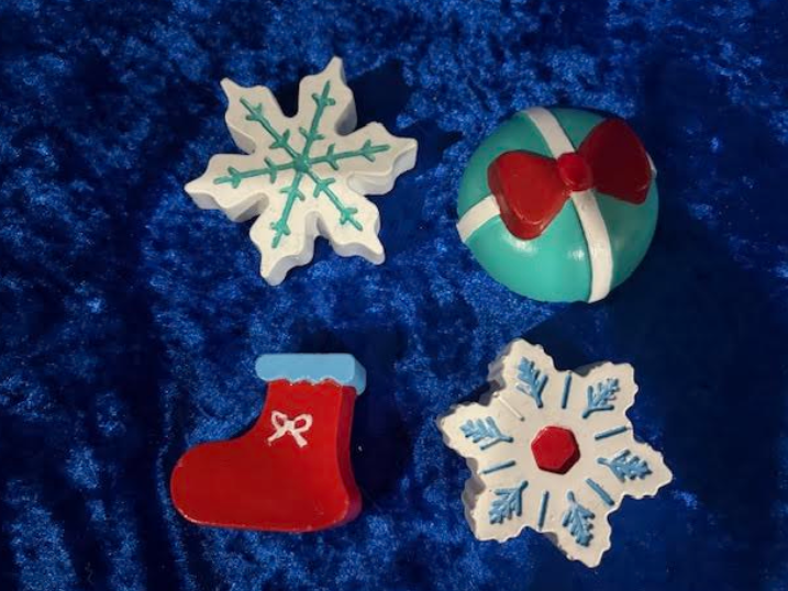 Four Christmas ornaments on blue fabric: two snowflakes, a red stocking, and a striped bauble with bow