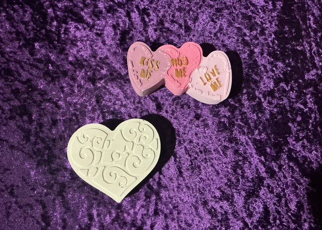 Three heart-shaped soaps, two pink and one cream, on a purple fuzzy background