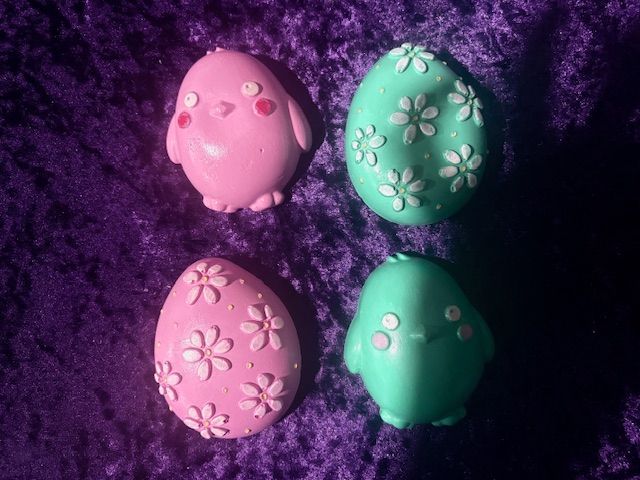 Four pastel decorated Easter eggs on purple fabric, pink and green with floral patterns and chick faces