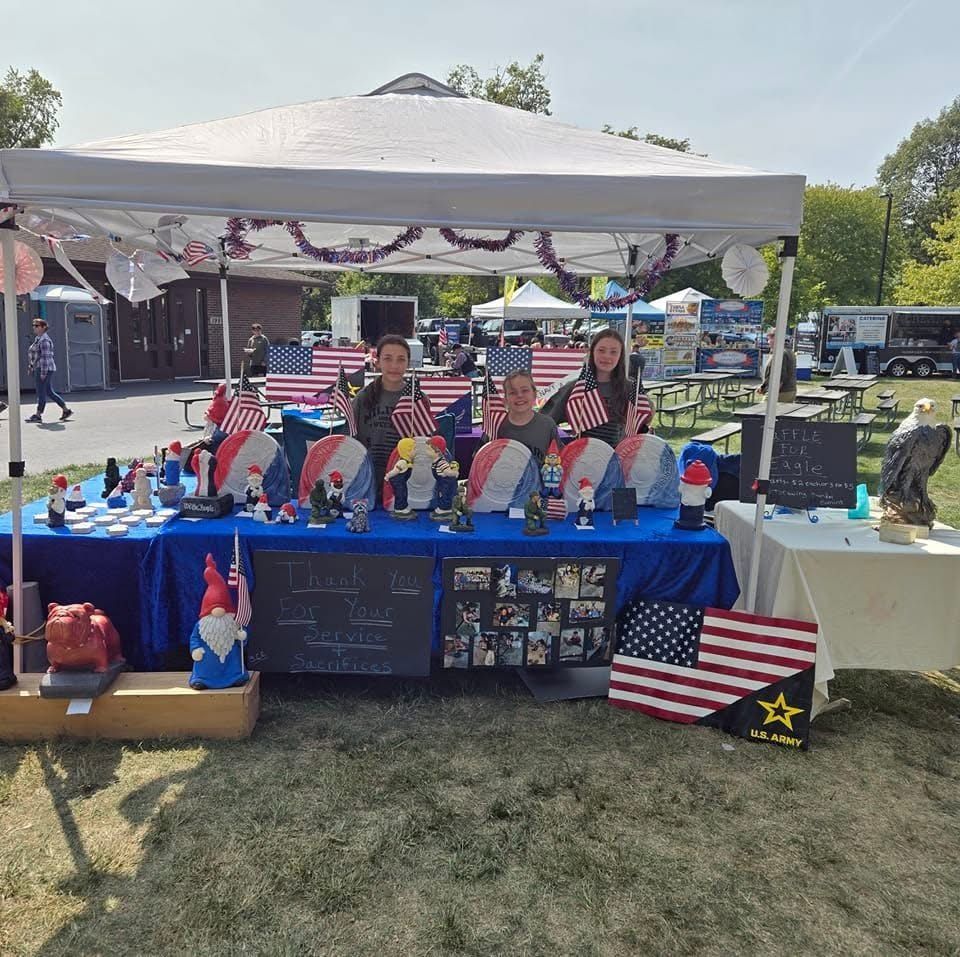 Outdoor booth with blue tablecloths, patriotic decorations, and two people behind a table at a park event