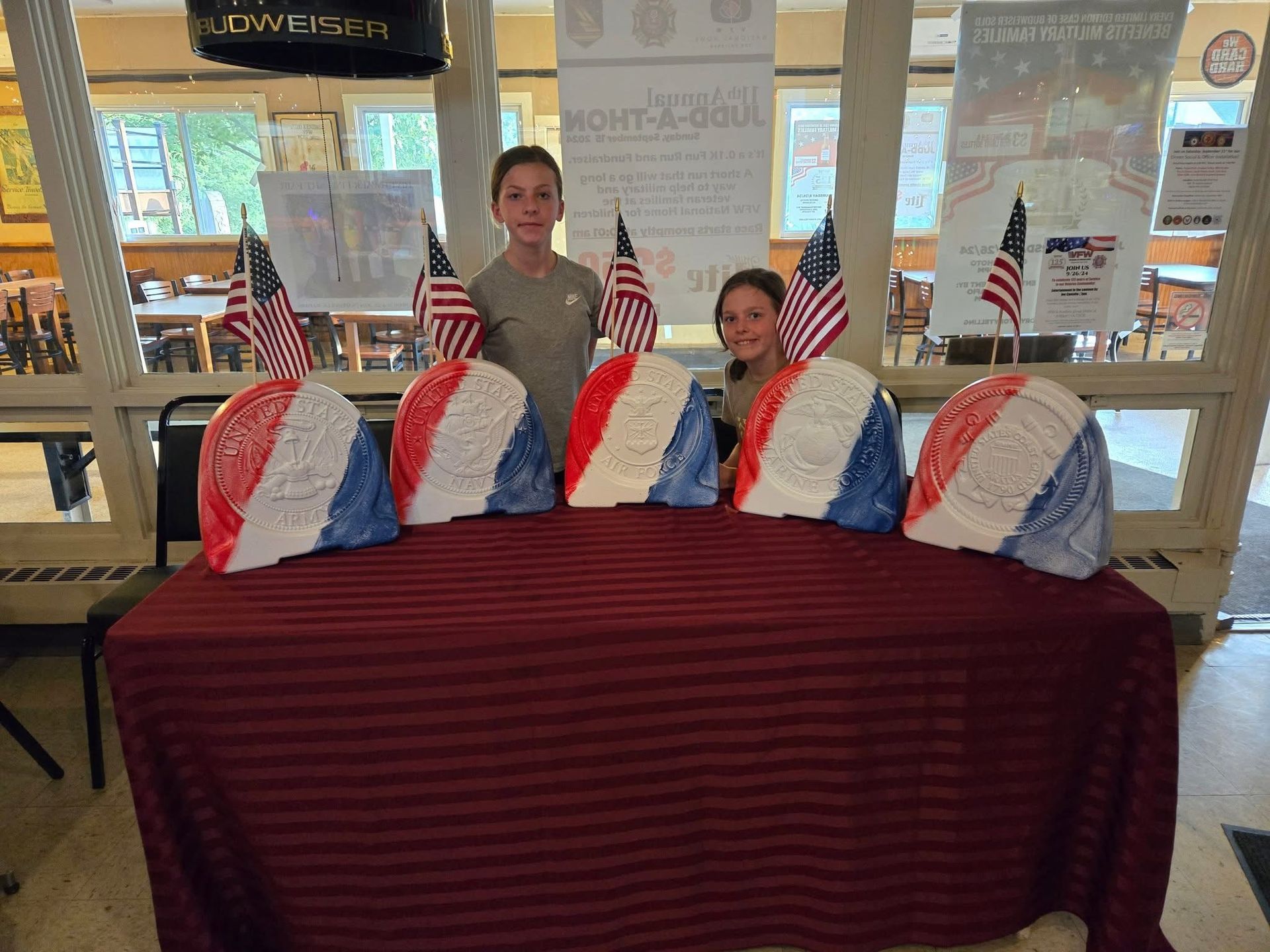 Two people behind a table draped in red, displaying red, white, and blue gift bags in a bright indoor space