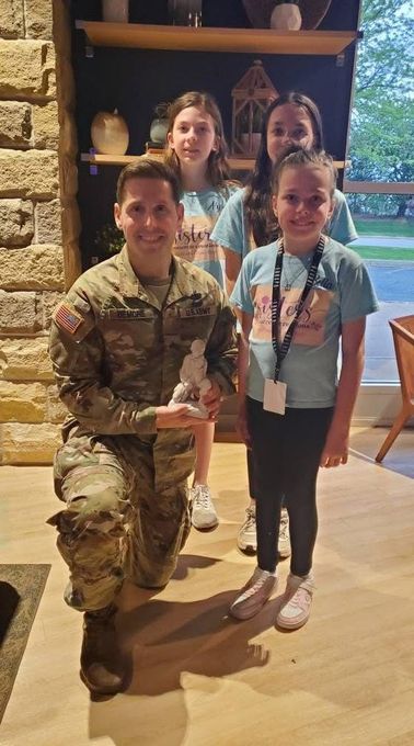 Soldier kneeling with three smiling children indoors, one holding a drink, near large windows and a stone fireplace