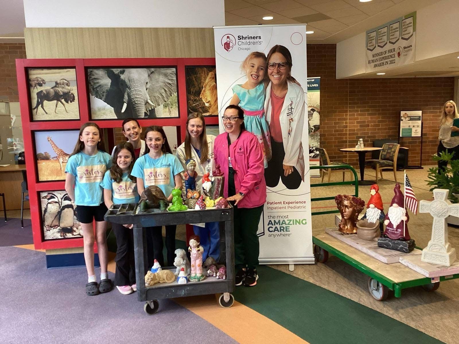 Group of children and adults at a display table in a school hallway beside a colorful poster and decorated carts